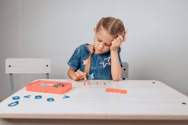 Portrait of a tired pensive girl sitting at a white table and solving intellectual problems placed on the table. Exhausted child. Childhood concept. photo with noise