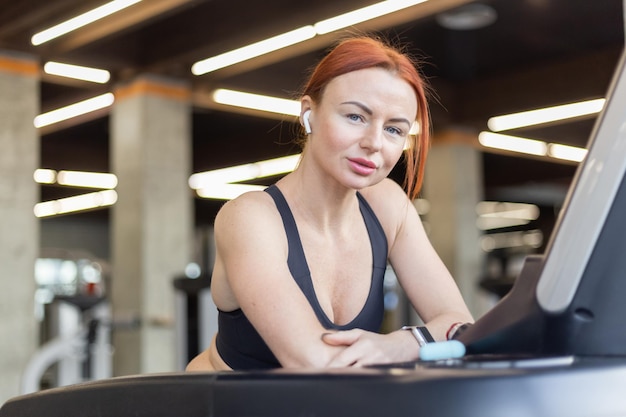 Ritratto di donna snella in forma dai capelli rossi stanca dietro il tapis roulant nella moderna palestra