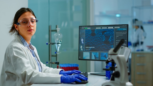 Photo portrait of tired scientist trying to smile at camera sitting in modern equipped laboratory typing on computer. chemist examining virus evolution using high tech for scientific research, vaccine