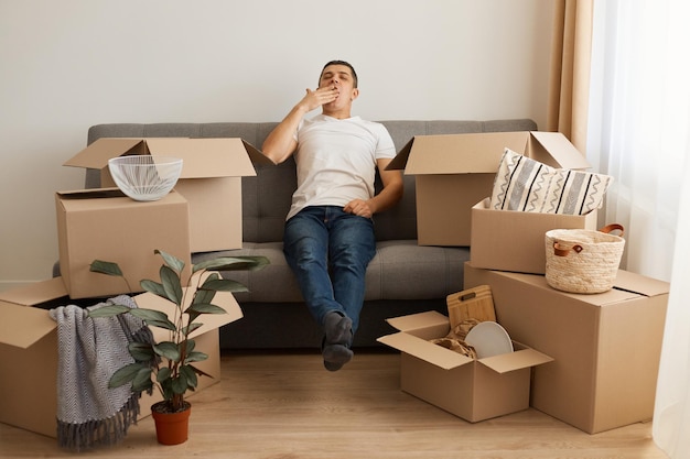 Портрет усталого сонного мужчины в белой повседневной футболке и джинсах, сидящего на диване, распаковывающего коробки после переезда, зевающего, прикрывающего рот ладонью, измученного распаковкой личных вещей.