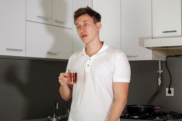 Portrait of tired and sleepy young man is drinking a tea in the morning in kitchen at home. He yawns and rubs his eyes, trying to wake up. Early morning on a weekday.