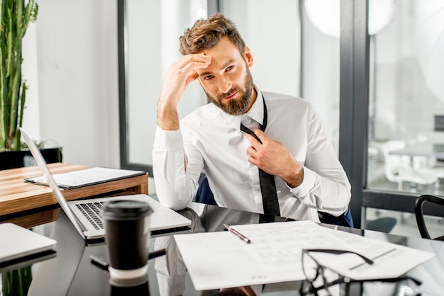 Portrait of a tired tax manager taking off a tie while working with tax documents and laptop at the office