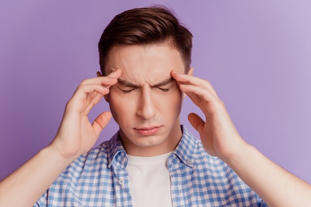 Портрет усталого нездорового парня, руки касаются головы, страдают от головной боли на фиолетовом фоне