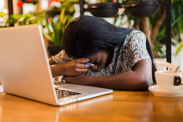 Portrait of a tired young african woman sitting at the table with laptop computer while sleeping at a cafe