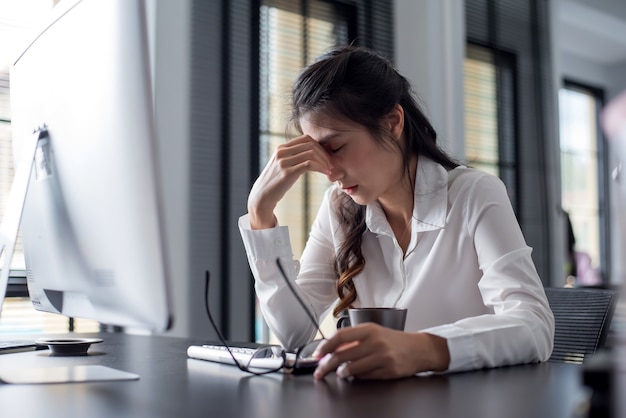 Portrait of a tired young asian businesswoman sitting at the table with laptop computer in office