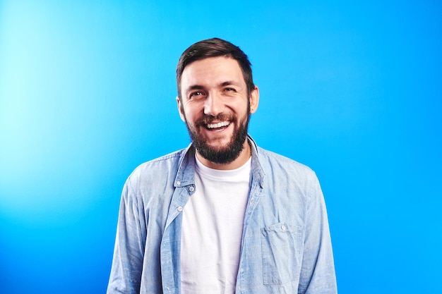 Portrait of toothy smiling and joyfully laughing handsome caucasian brutal bearded man dressed in casual isolated on blue background