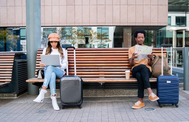 Portrait of a tourist couple sitting on bench outside airport or train station. Woman using laptop and man looking at map