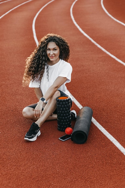 Portrait of a trainer on a stadium. She is sitting near mfr roller, yoga mat and her phone.