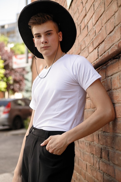 Ritratto di un giovane alla moda in città. primo piano ritratto ragazzo modello in un cappello nero uno sfondo di muro di mattoni