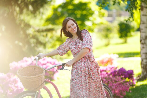 Portrait of trendy young woman in long pink floral dress stop to riding on vintage bike with basket for purchases on flowers background outdoors. Pretty female recreation time in spring or summer park