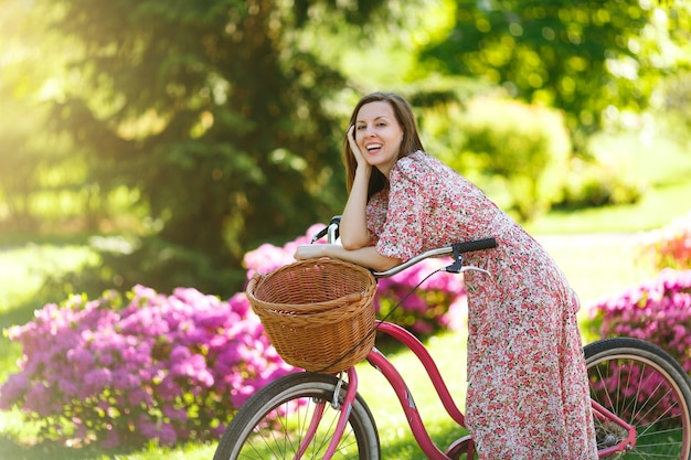 Portrait of trendy young woman in long pink floral dress stop to riding on vintage bike with basket for purchases on flowers background outdoors. Pretty female recreation time in spring or summer park