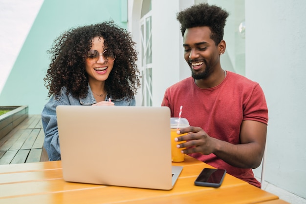 Ritratto di due amici afro utilizzando laptop mentre beve succo di frutta fresca all'aperto in una caffetteria.