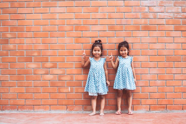Portrait of two asian kid girls pointing up on brick wall background