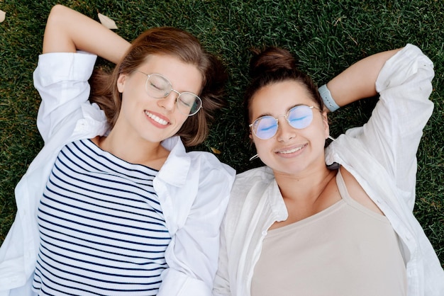 Portrait of two attractive female students wearing stylish glasses lying on the green grass