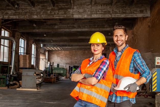 Портрет двух привлекательных улыбающихся фабричных рабочих, мужчины и женщины в оранжевых защитных жилетах и серых перчатках в помещении на деревообрабатывающем производстве Оборудование и техника на заднем плане