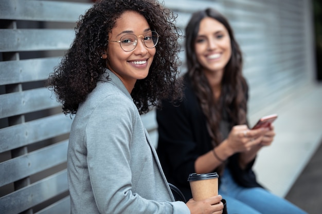 Portrait of two beautiful businesswomen using smart phone while looking at camera sitting on a bench on the street.