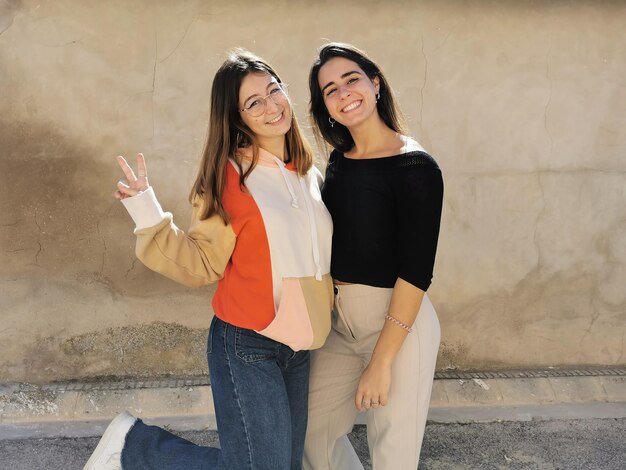 Photo portrait of two beautiful girl friends embracing each other making the victory sign