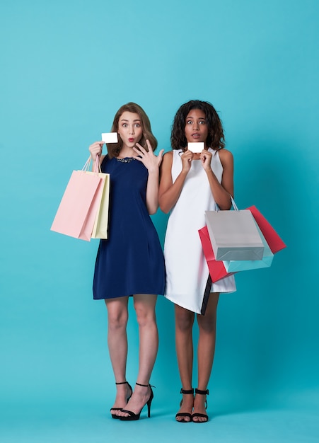 Photo portrait of two beautiful young woman  showing credit card and shopping bag isolated over blue background.