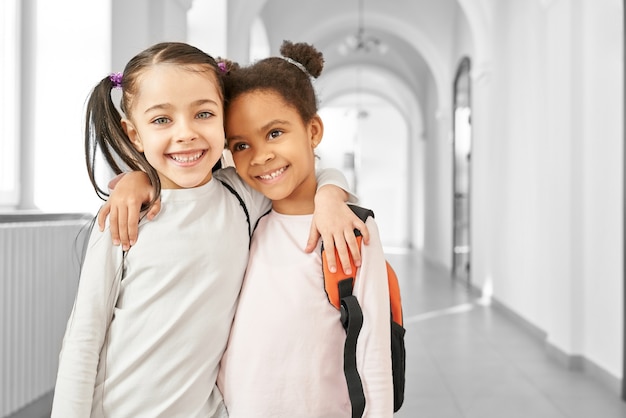 Portrait of two best friends little school girls standing at corridor, embracing each other and smiling.