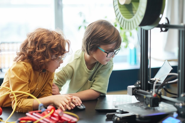 Portrait of two boys using 3D printer during engineering and robotics class at school, copy space