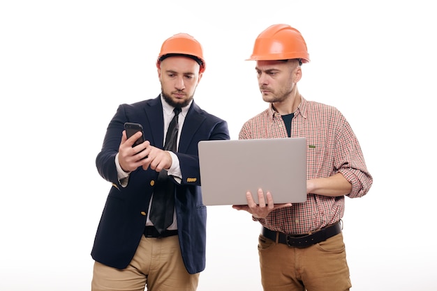 Portrait of two builders in protective orange helmets standing on white isolated background and looking at laptop display. Discuss construction project