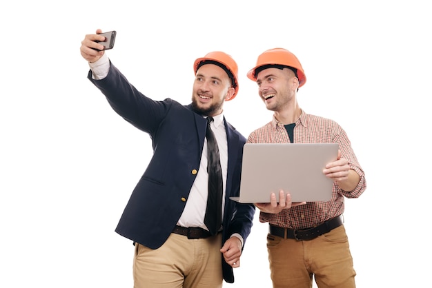 Photo portrait of two builders in protective orange helmets standing on white isolated background and looking at laptop display. discuss construction project