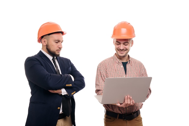 Foto ritratto di due costruttori in caschi protettivi arancioni in piedi su sfondo bianco isolato e guardando il display del laptop. discuti il progetto di costruzione