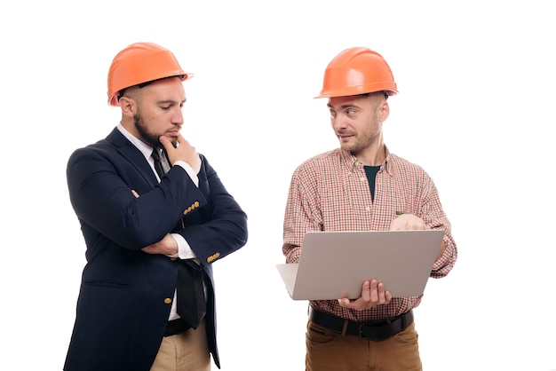 Photo portrait of two builders in protective orange helmets standing on white isolated background and looking at laptop display. discuss construction project