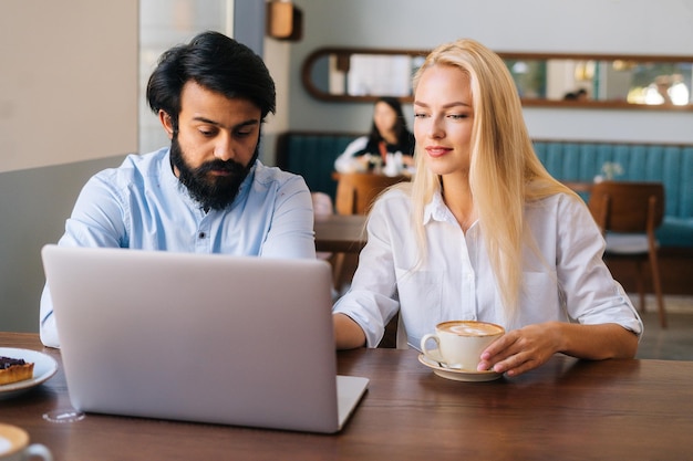 Portrait of two business people of bearded young businessman and pretty Caucasian blonde woman working with laptop in cafe and drinking coffee Friendly colleagues discussing project in coffee shop