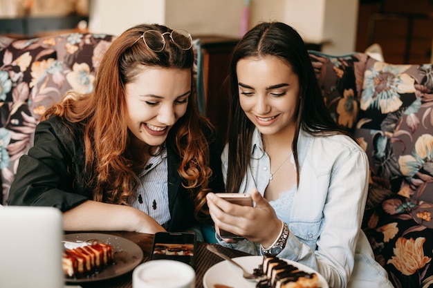 Photo portrait of two cheerful friend looking to a smartphone screen smiling while sitting in a cafe eating cheesecake and drinking coffee.