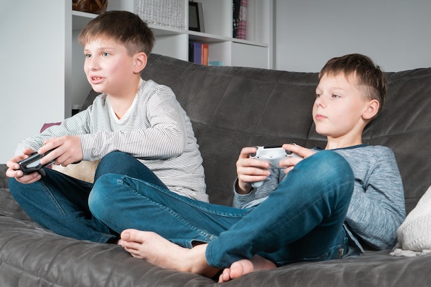 Portrait of two concentrated teenage boys sitting on grey sofa at home holding gaming joystick playing videogames