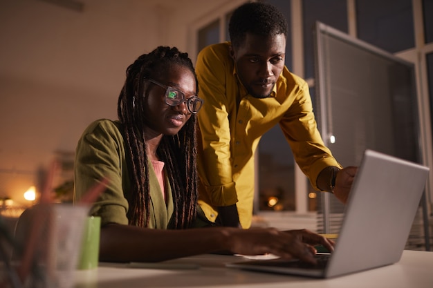 Портрет двух современных афроамериканцев, смотрящих на экран ноутбука допоздна в тусклом офисе