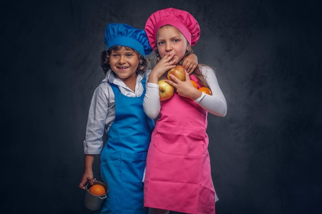 Portrait of two cute little cooks. Little boy with brown curly hair dressed in a blue cook uniform and beautiful schoolgirl dressed in a pink cook uniform, cuddling together while holds cookware and f