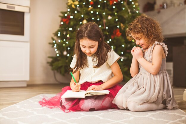 Portrait of two cute little sisters writing a letter to santa on a floor under christmas tree with lights at their house kids believe in magic concept