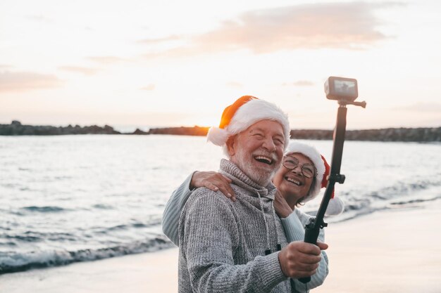 Portrait of two cute old persons having fun and enjoying together at the beach on christmas days at the beach wearing Christmas hats Looking and holding a camera taking videos of vacations