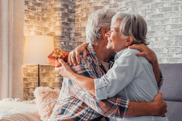 Portrait of two cute and old seniors at home having fun\
together mature man giving a gift at his wife for christmas or\
anniversary surprised pensioner woman hugging his husbandxa