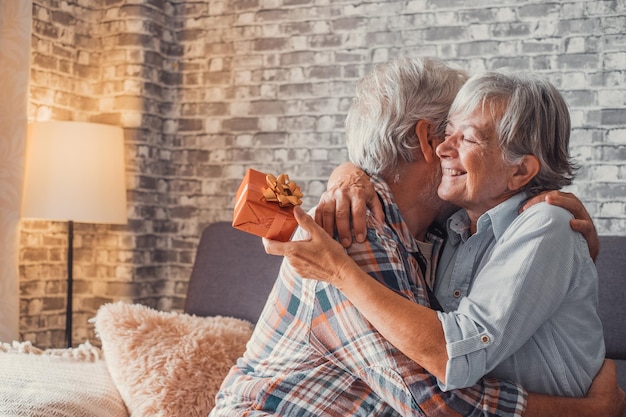Portrait of two cute and old seniors at home having fun\
together mature man giving a gift at his wife for christmas or\
anniversary surprised pensioner woman hugging his husbandxa