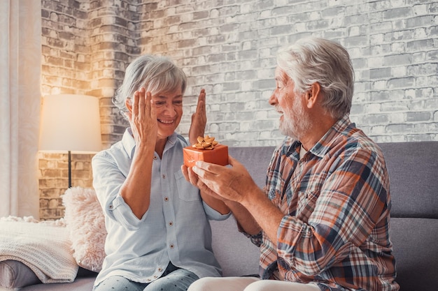 Portrait of two cute and old seniors at home having fun together Mature man giving a gift at his wife for Christmas or anniversary Surprised pensioner woman looking at the present