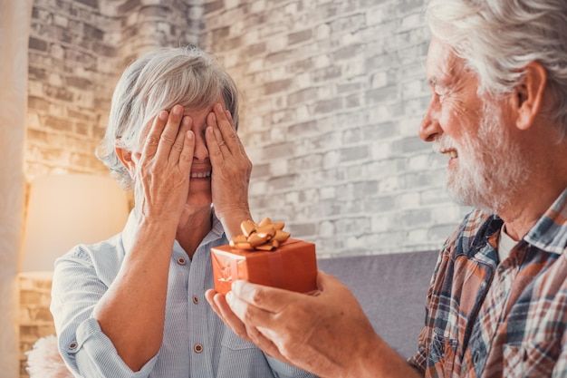 Portrait of two cute and old seniors at home having fun\
together mature man giving a gift at his wife for christmas or\
anniversary surprised pensioner woman looking at the presentxa