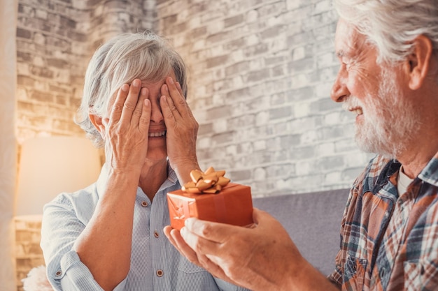 Portrait of two cute and old seniors at home having fun\
together mature man giving a gift at his wife for christmas or\
anniversary surprised pensioner woman looking at the presentxa