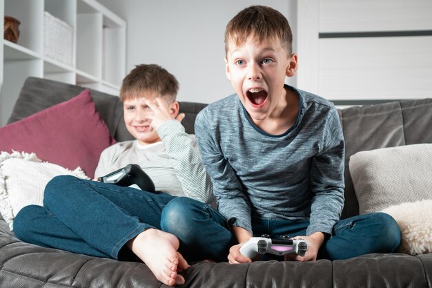 Portrait of two excited teenage boys sitting at home holding gaming controller joystick playing videogames shouting