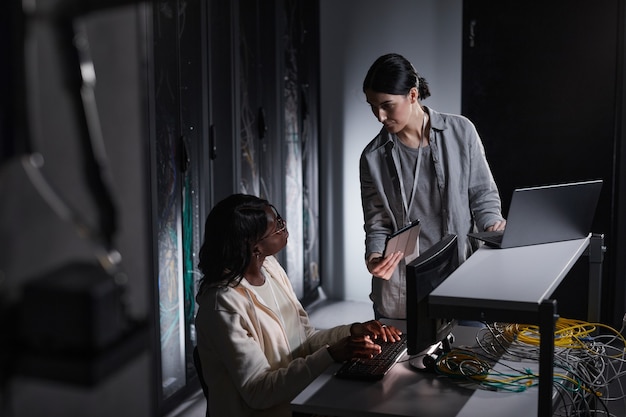 Портрет двух сетевых инженеров женского пола, использующих ноутбук во время совместной работы в темной серверной комнате, копировальное пространство
