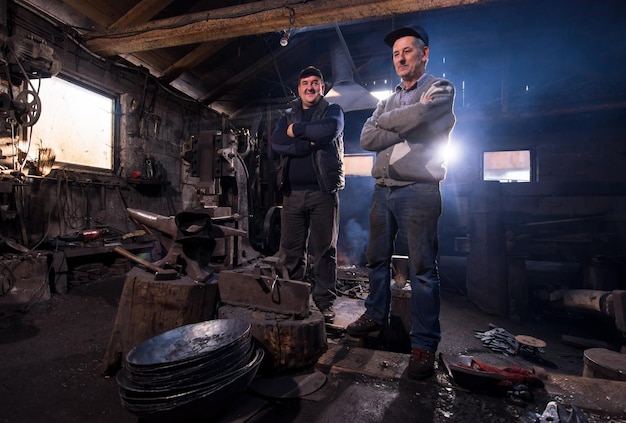 portrait of two generations confident blacksmith standing with crossed arms in the traditional workshop