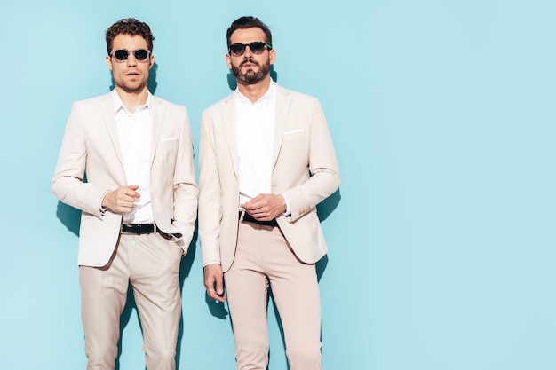 Portrait of two handsome confident stylish hipster lambersexual models Sexy modern men dressed in white elegant suit Fashion male posing in studio near blue wall