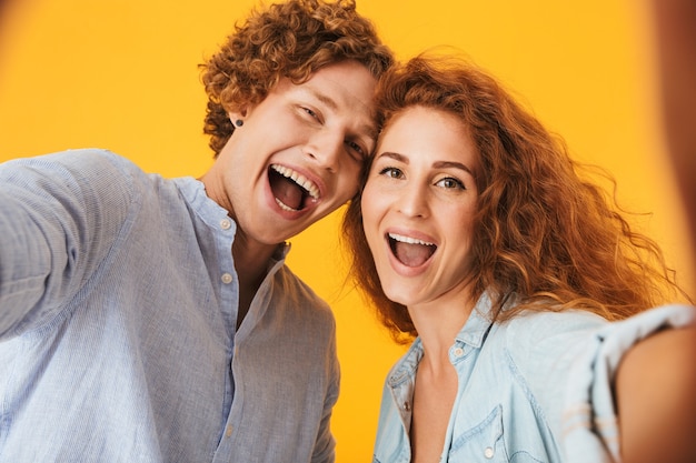 Portrait of two happy people man and woman laughing and taking selfie photo, isolated over yellow background