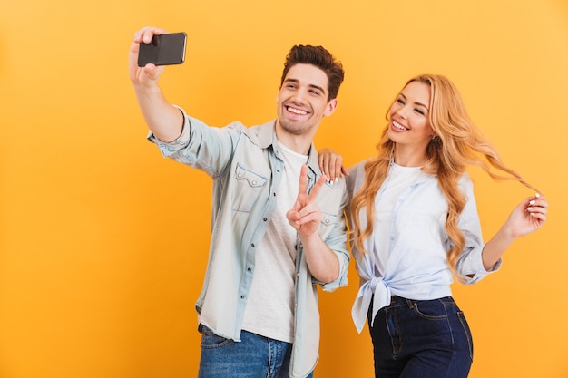 Portrait of two happy people man and woman taking selfie photo on smartphone while showing peace sign, isolated over yellow wall