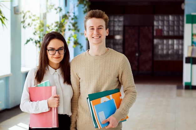 Портрет двух учеников средней школы с учебниками, учебниками и папками перед занятиями в школьном коридоре в солнечный день