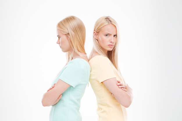 Photo portrait of two indignant girls standing back to each other with arms crossed isolated over white wall
