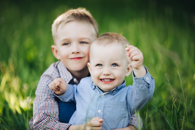 Portrait of two little stylish happy brother with beautiful blue eyes playing in park embracing and looking at the camera Boys wearing in shirts posing against green grass background