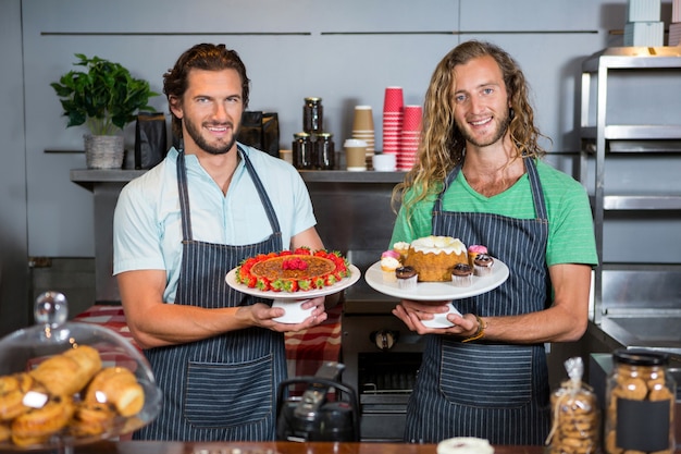 Portrait of two male staffs holding dessert on cake stand at counter